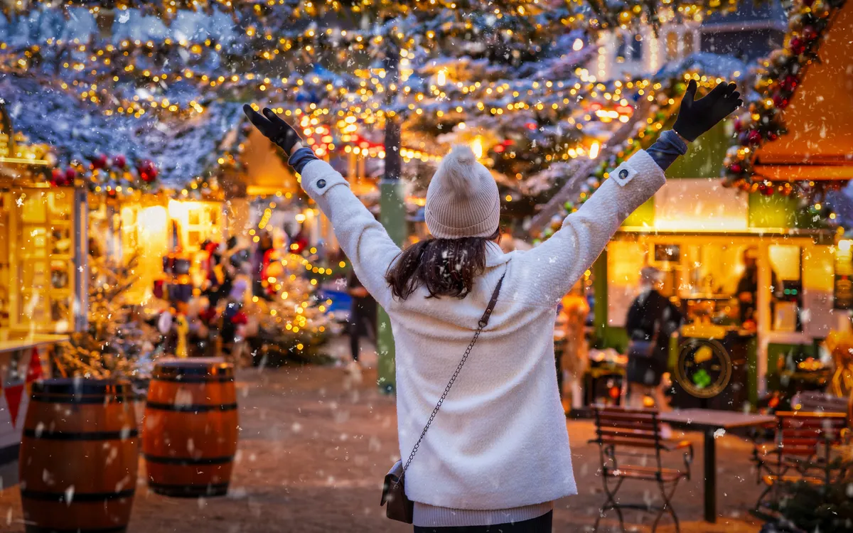 Person auf Weihnachtsmarkt, Hände hoch, umgeben von Lichtern und Dekorationen.