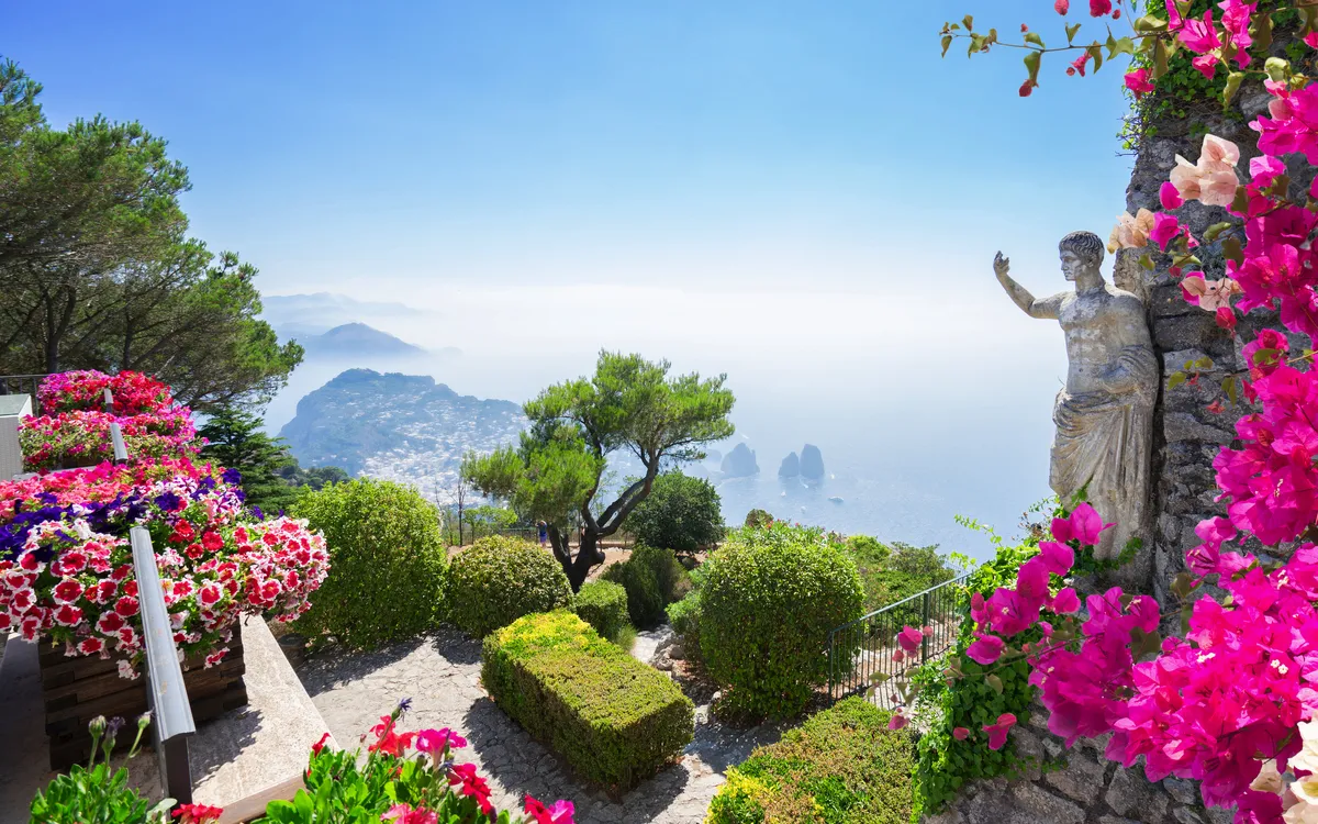 Blumenreicher Garten mit Blick auf das Meer und eine Statue auf einem sonnigen Tag.