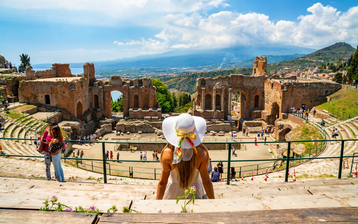 Frau mit Hut sitzt vor antikem römischen Theater mit Blick auf Landschaft.