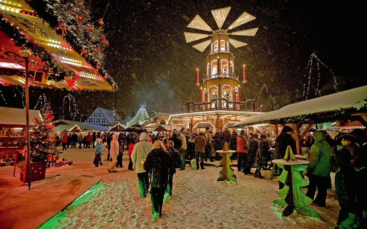 Menschenmenge auf beleuchtetem Weihnachtsmarkt bei Nacht, Schnee fällt.
