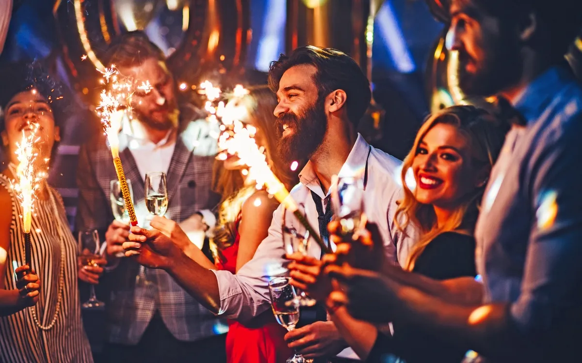 Gruppe von Menschen feiert fröhlich auf einer Party mit Wunderkerzen und Getränken in der Hand.