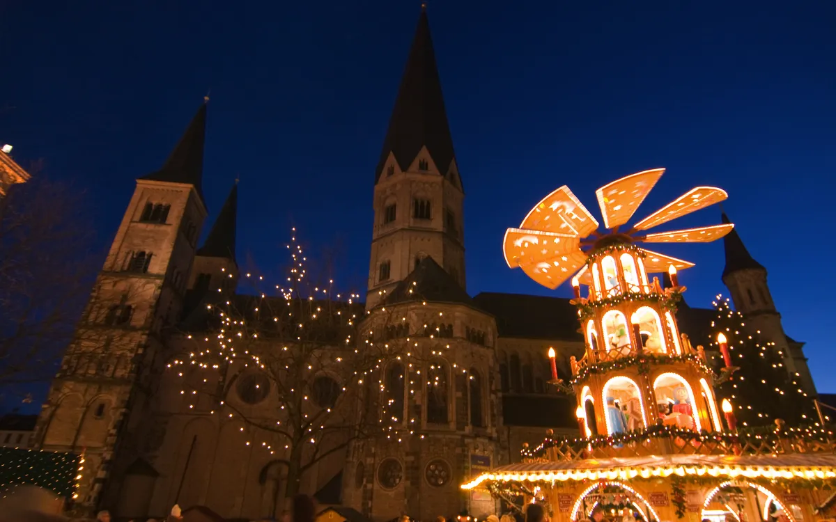 Weihnachtsmarkt mit beleuchteter Weihnachtspyramide vor einer Kirche bei Nacht im Hintergrund.