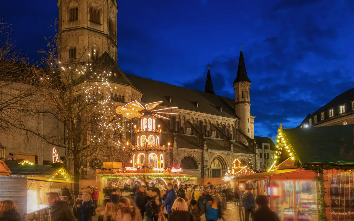 Menschen auf einem beleuchteten Weihnachtsmarkt vor historischer Kirche am Abend.
