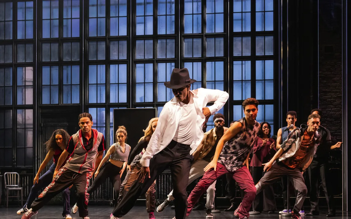Tänzer in dynamischer Choreografie auf einer Bühne mit großen Fenstern im Hintergrund.
