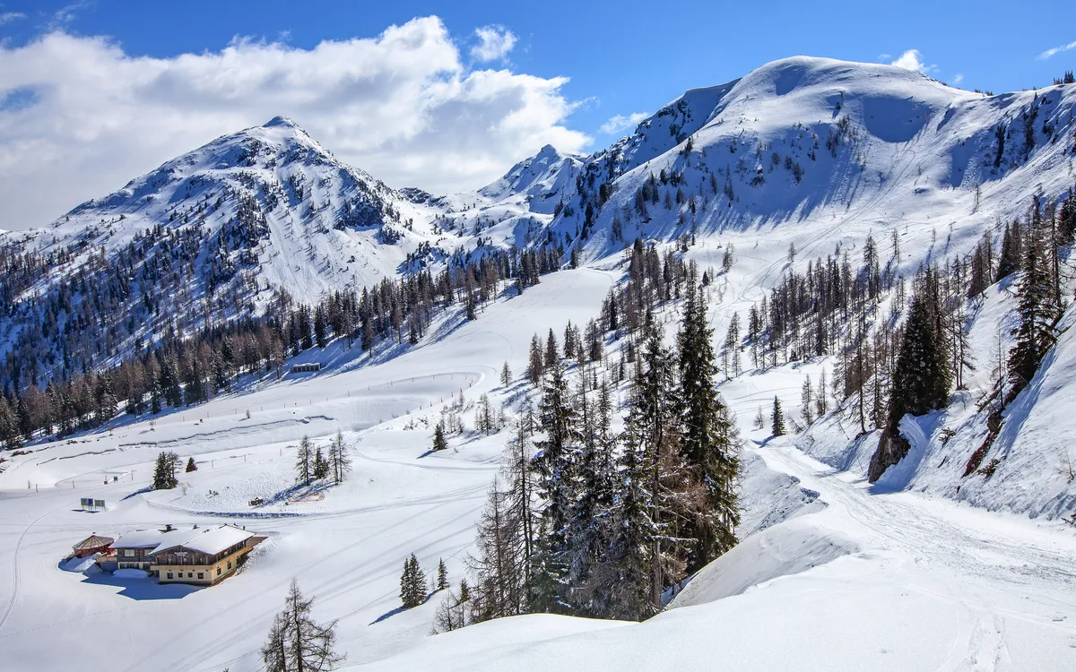 Verschneite Berglandschaft mit Hütte und Kiefern unter blauem Himmel.