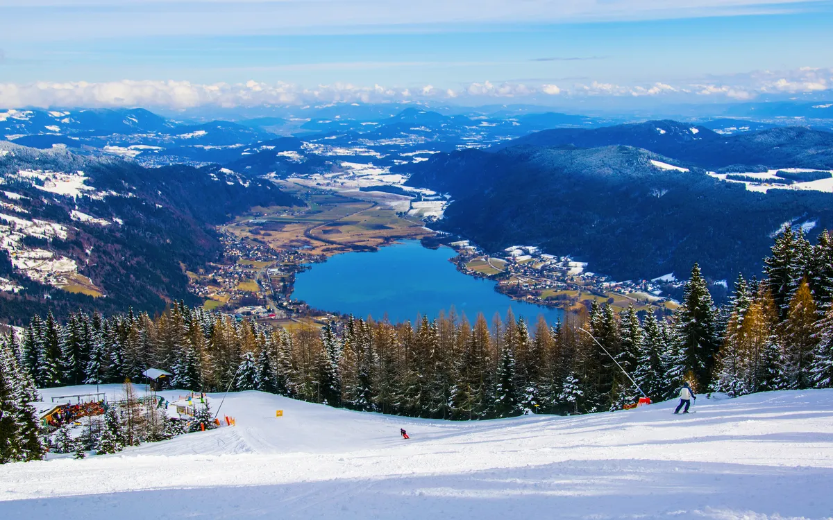 Skifahrer auf schneebedeckter Piste mit Blick auf einen See und umgebende Berge.