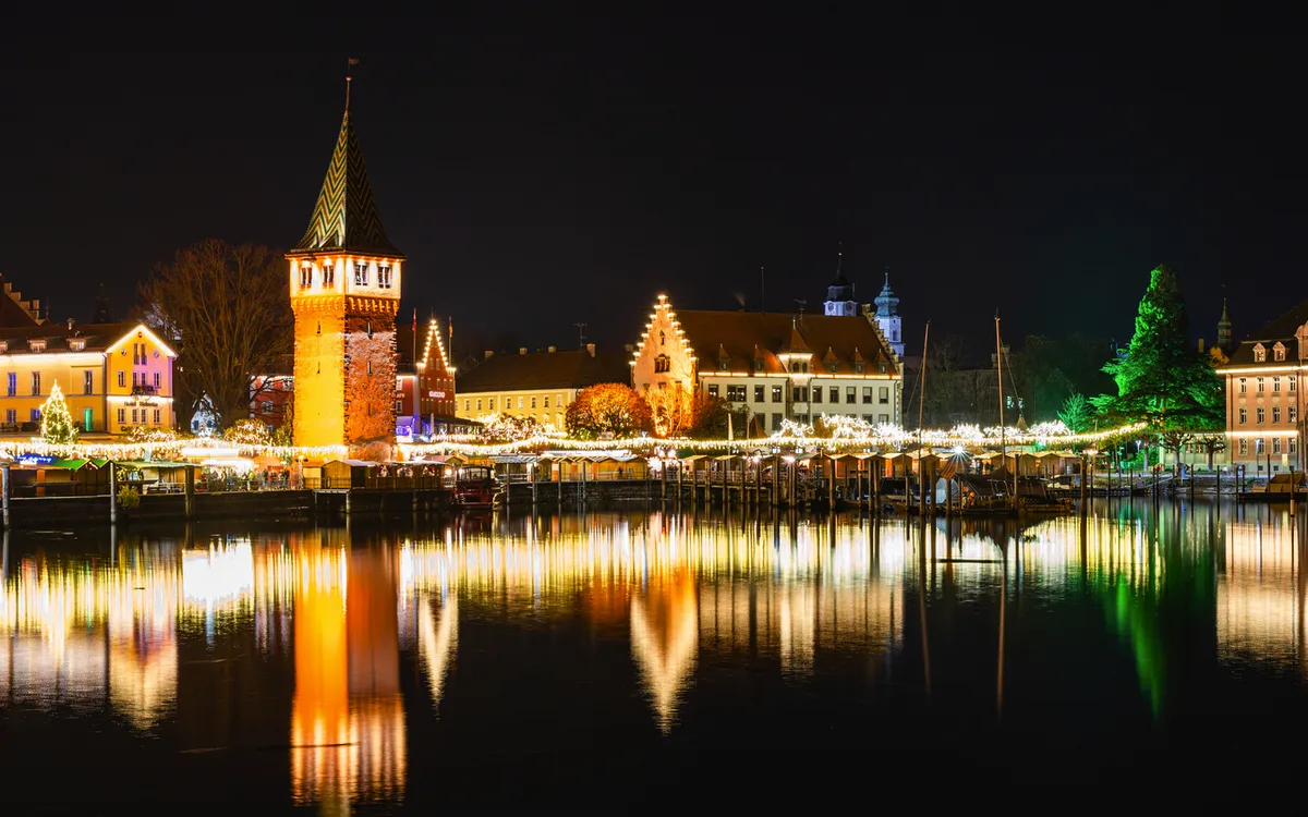 Hafenstadtansicht bei Nacht, beleuchtete Gebäude spiegeln sich im Wasser.