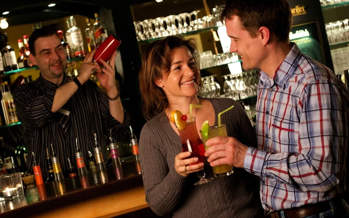 Drei Personen an einer Hotelbar, zwei trinken Cocktails, Barkeeper im Hintergrund.