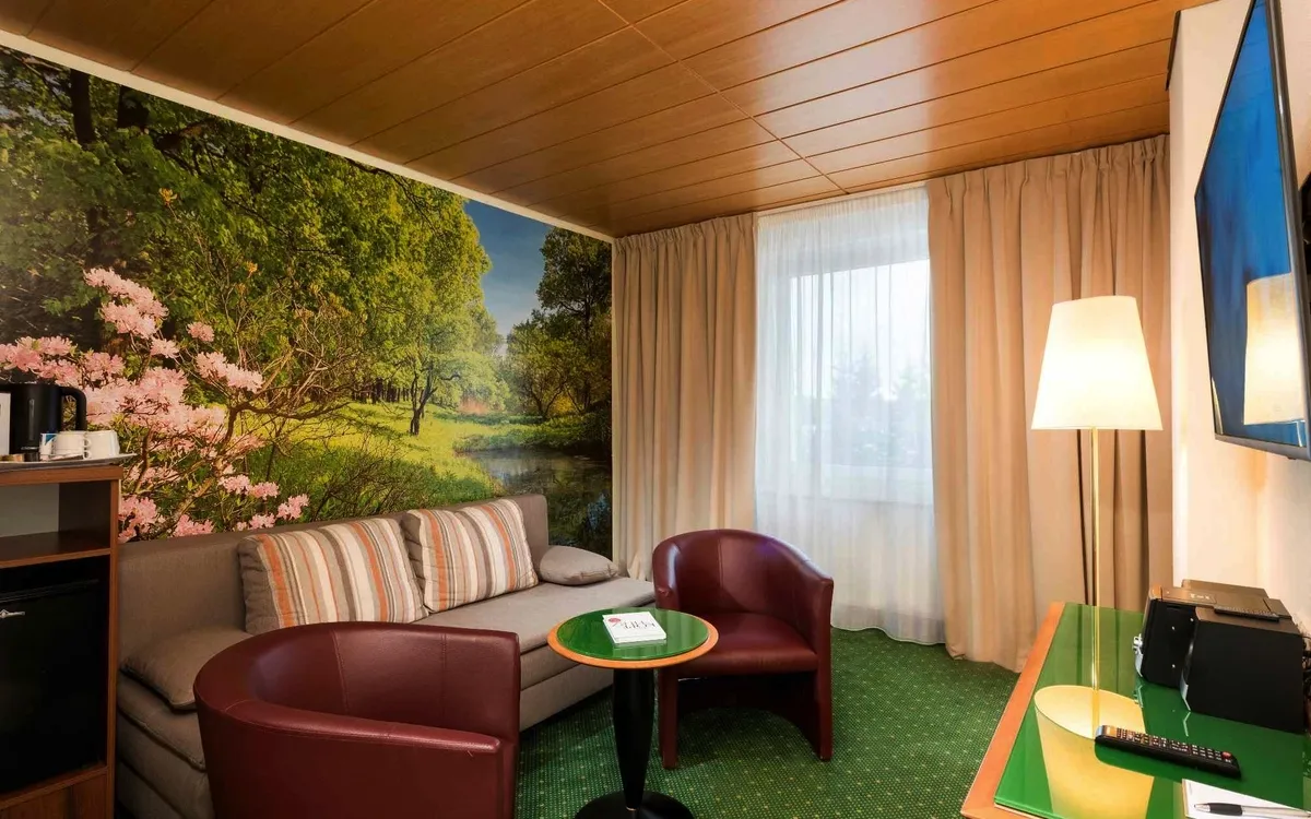 Hotelzimmer mit Sofa, Tisch, zwei Sesseln und Wandbild einer Waldlandschaft.