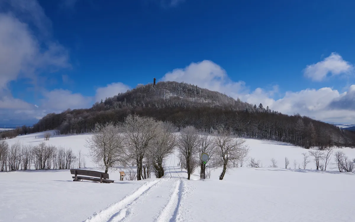 Verschneite Landschaft mit Hügel, Wald und blauem Himmel im Hintergrund.