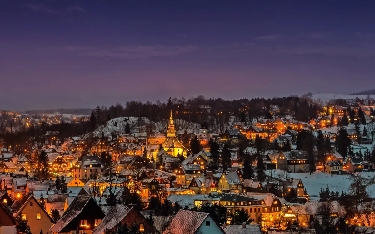 Verschneite Stadtlandschaft bei Nacht mit beleuchteten Häusern und Kirche.