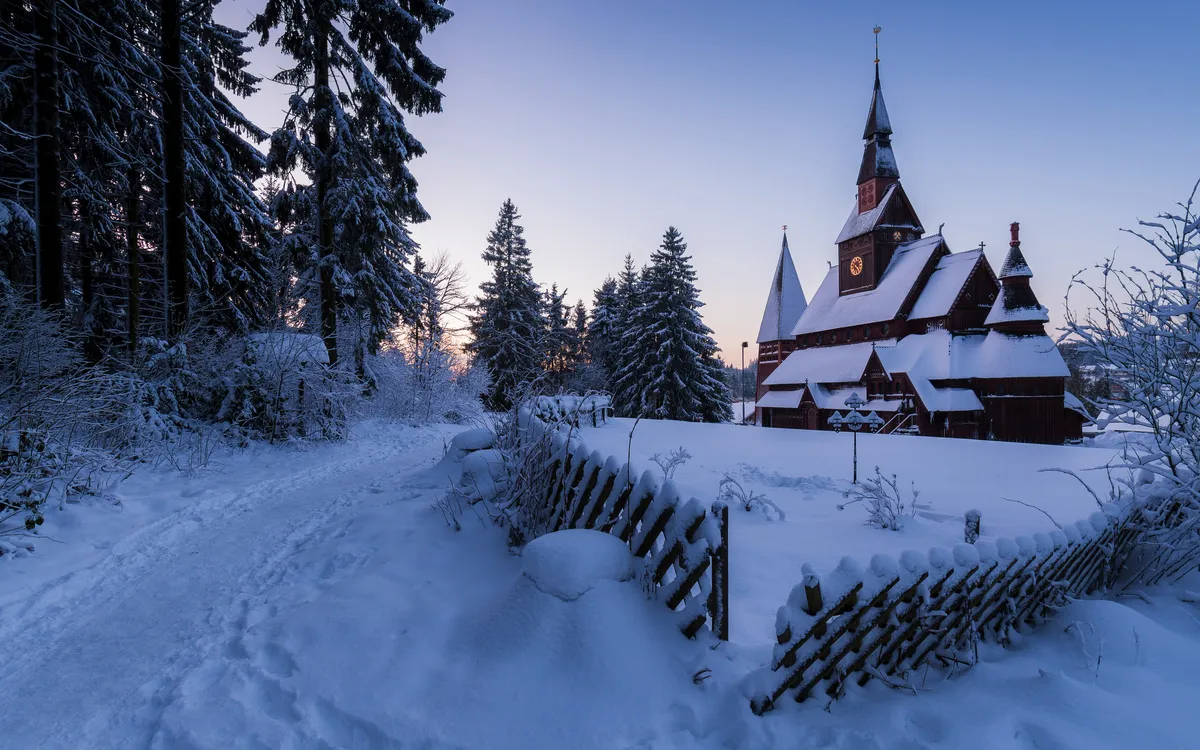 Winterlandschaft mit verschneiter Holzkapelle und Tannenbäumen im Hintergrund.