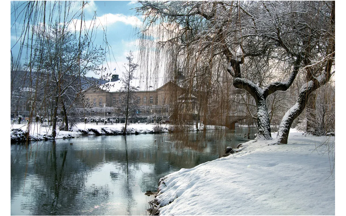 Verschneite Flusslandschaft in Bad Kissingen im Winter, alte Gebäude im Hintergrund.