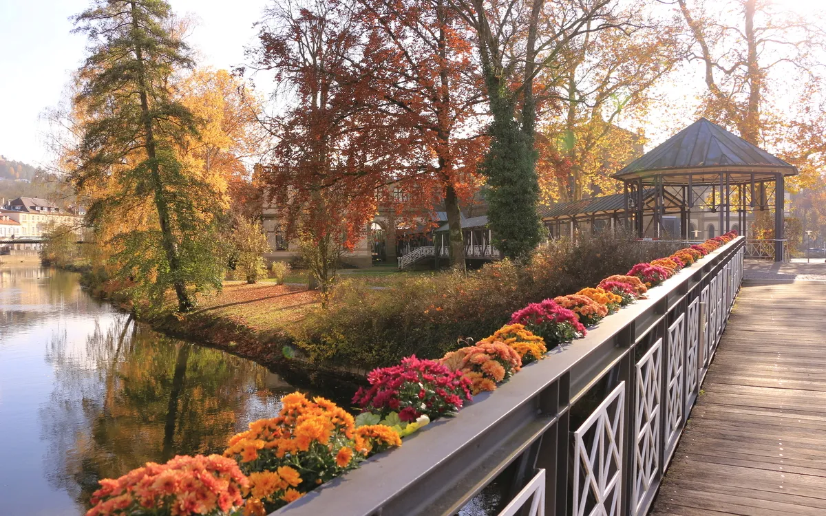 Brücke mit bunten Blumen, Herbstbäume im Hintergrund, See und Pavillon.