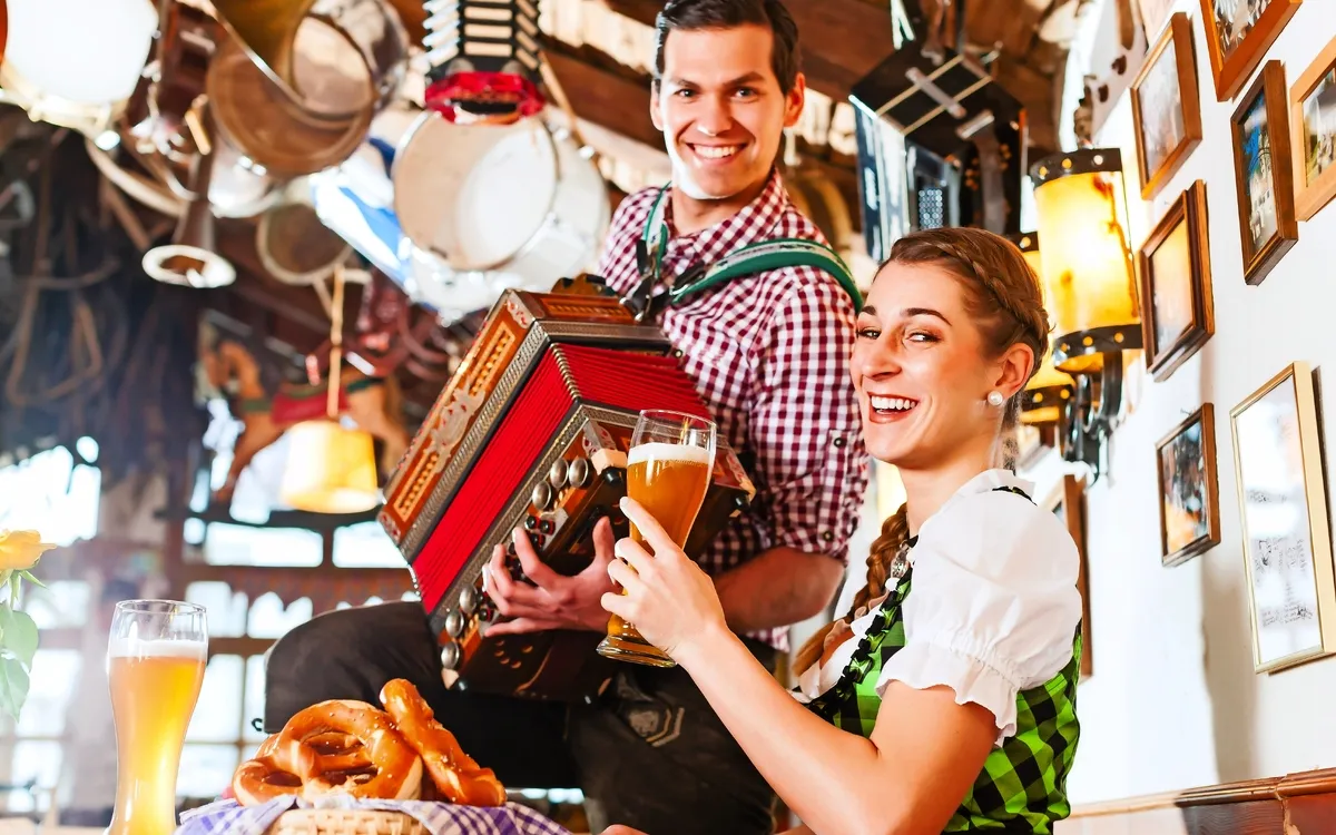 Zwei Personen in traditioneller Kleidung in einer bayerischen Wirtschaft mit Bier und Brezeln.