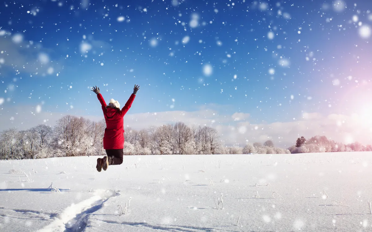 Person springt im Schnee, rote Jacke, sonniger Wintertag, Schneeflocken fallen