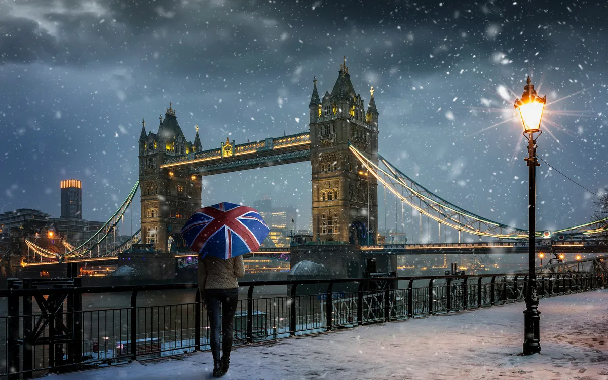 Tourist mit Regenschirm mit britischer Flagge steht bei Schneefall vor der Tower Bridge in London.