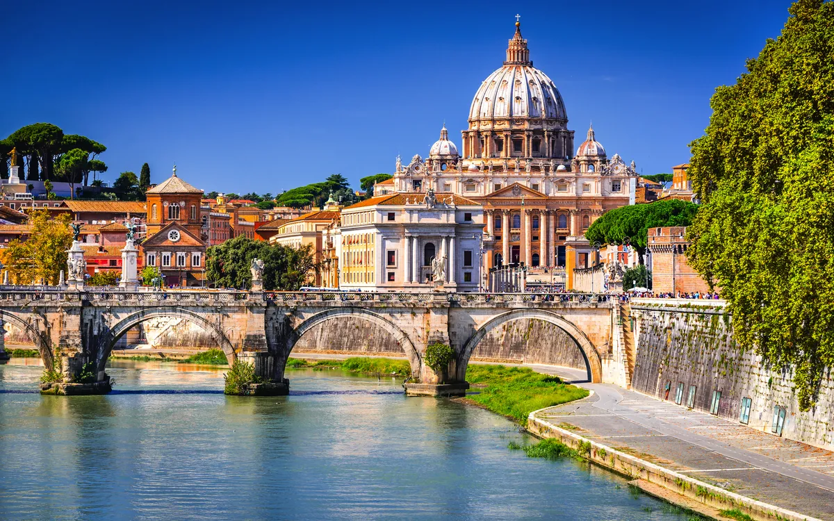 Der Petersdom in Rom mit der Engelsbrücke und dem Fluss Tiber im Vordergrund.