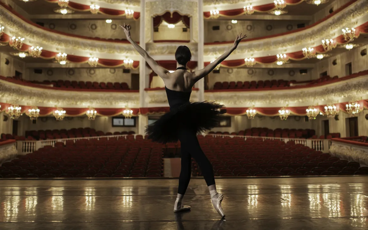 Balletttänzerin auf leerer Theaterbühne vor Publikumssitzen und Kronleuchtern.