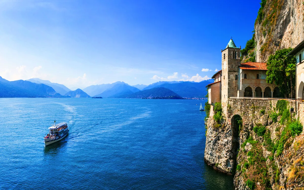 Kloster am Lago Maggiore mit Bergblick und vorbeifahrendem Boot.