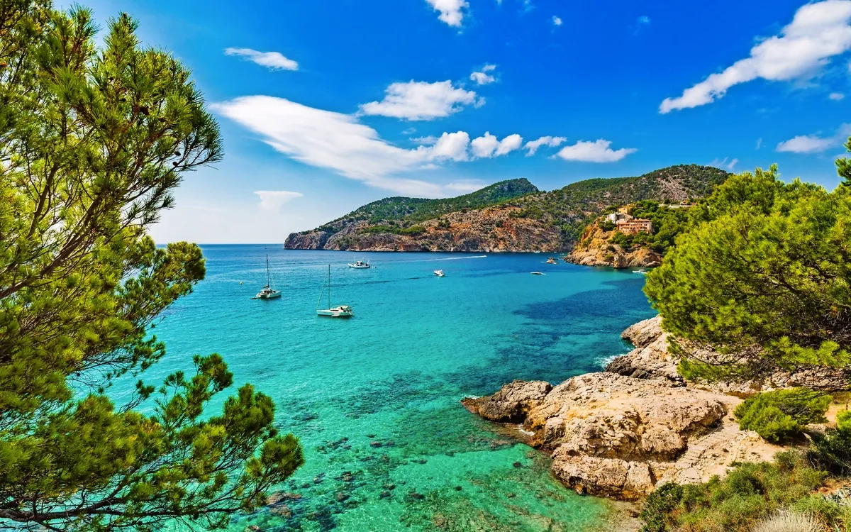 Küstenlandschaft mit blauem Meer, Segelbooten und grünen Bäumen unter blauem Himmel.