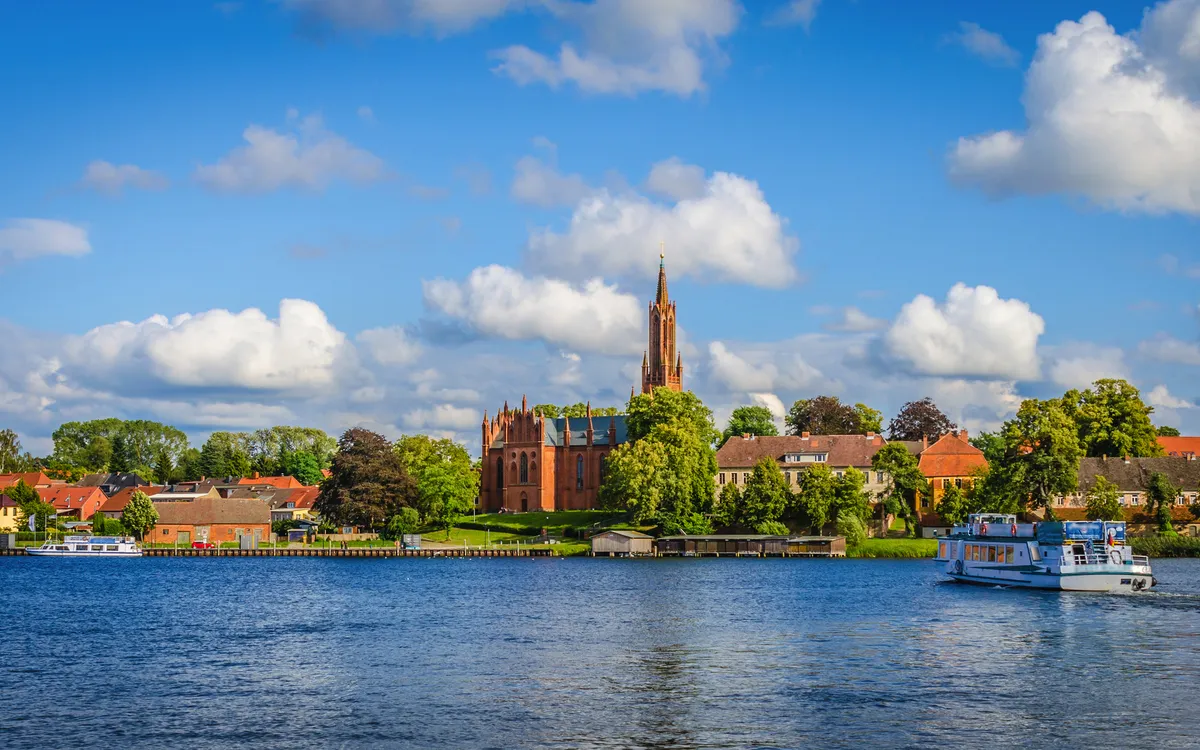 Kleinstadt mit Kirche und Boot auf Fluss, blauer Himmel, weiße Wolken.