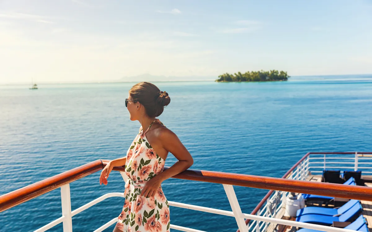 Frau in Sommerkleid steht auf Kreuzfahrtschiff, schaut aufs Meer und eine kleine Insel