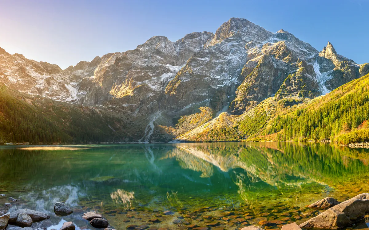 Bergsee mit klaren Wasser, umgeben von bewaldeten Bergen und strahlendem Sonnenlicht.