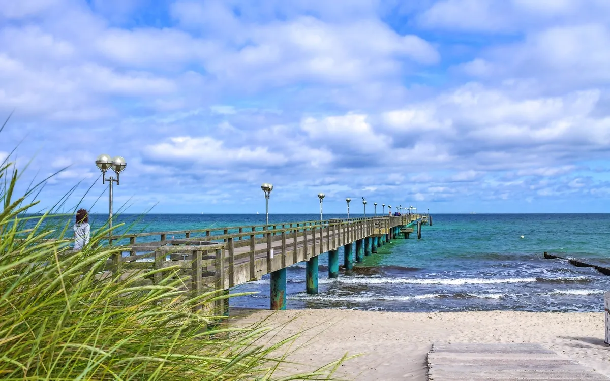Langer Holzsteg führt über Sandstrand zum Meer unter bewölktem Himmel.