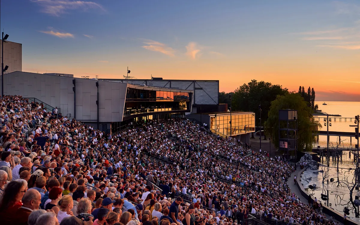 Menschenmenge sitzt bei Sonnenuntergang in einem Freilufttheater am Wasser.