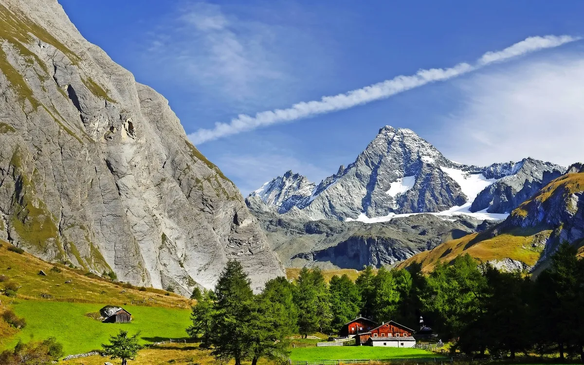 Berglandschaft mit grüner Wiese, Bäumen und Almhütte vor schneebedeckten Gipfeln.