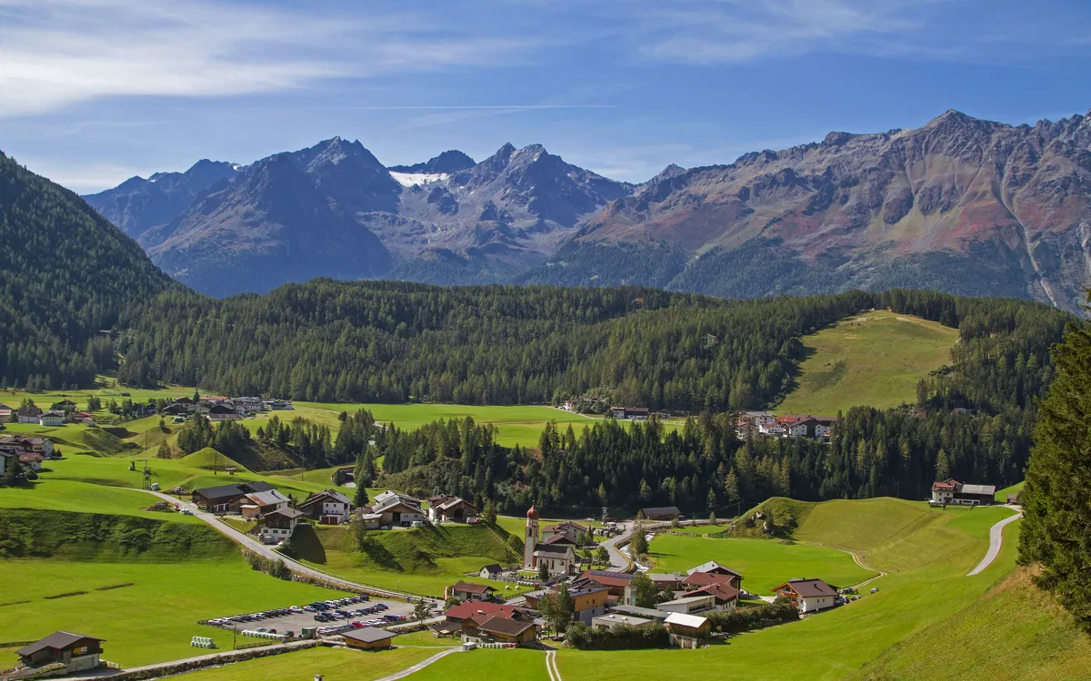Bergdorf in grüner Alpenlandschaft mit Häusern und umliegenden Bergen bei klarem Himmel.