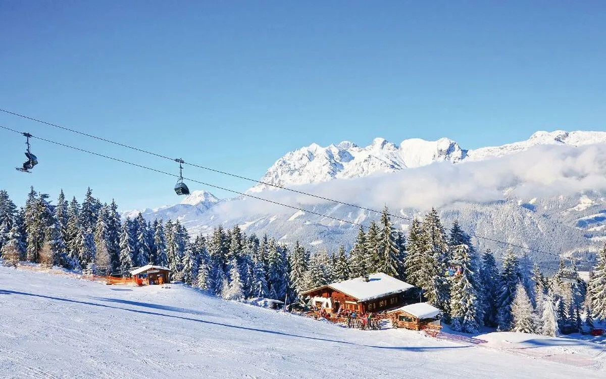 Verschneite Alpenlandschaft mit Skihütte, Bäumen und Skilift bei blauem Himmel.