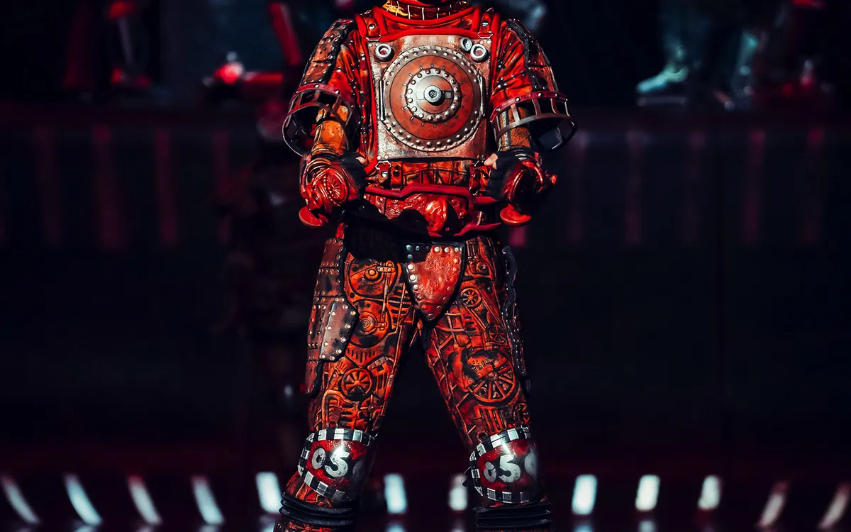 Person in futuristischem, rustfarbenem Kostüm auf einer Bühne, umgeben von weiteren Gestalten.