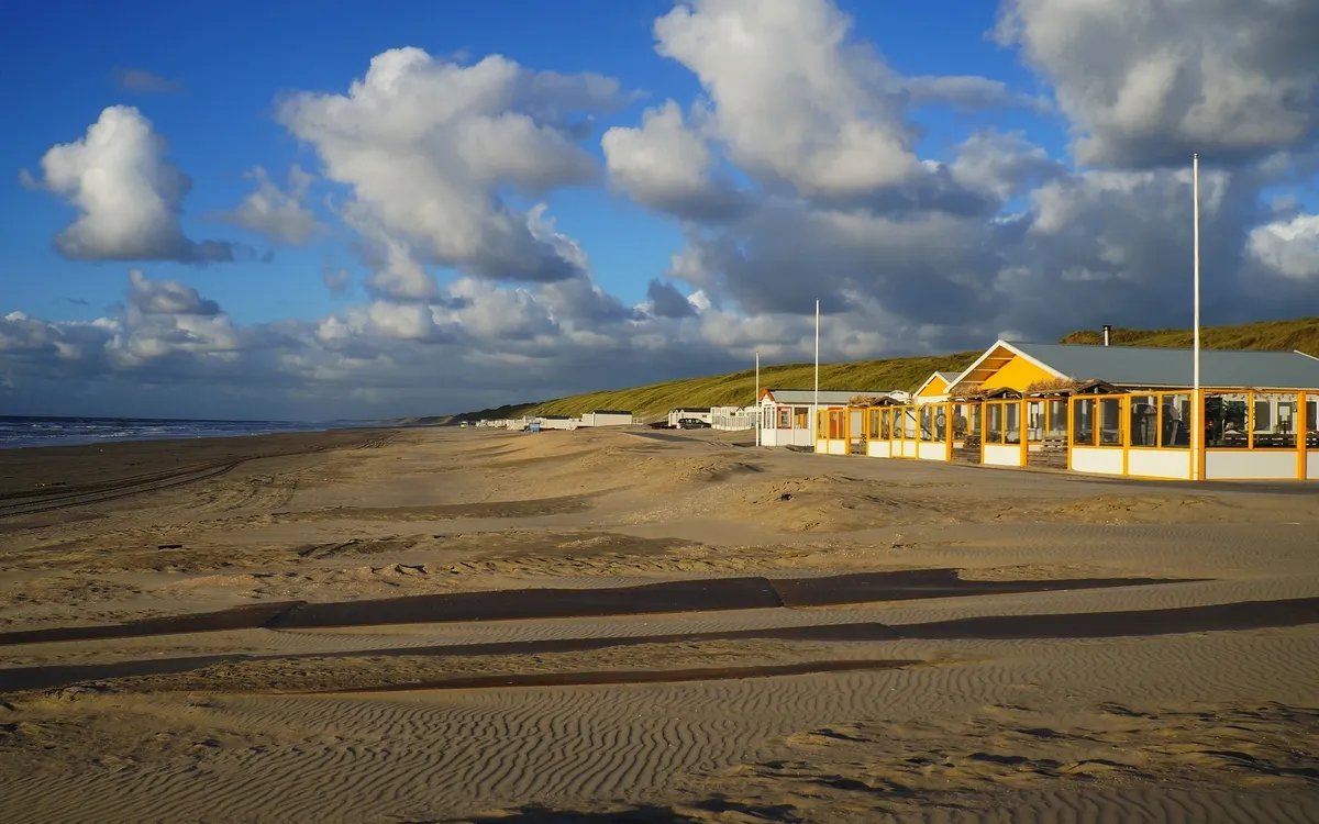 Strand mit Sanddünen und Ferienhäusern unter blauem Himmel mit Wolken.