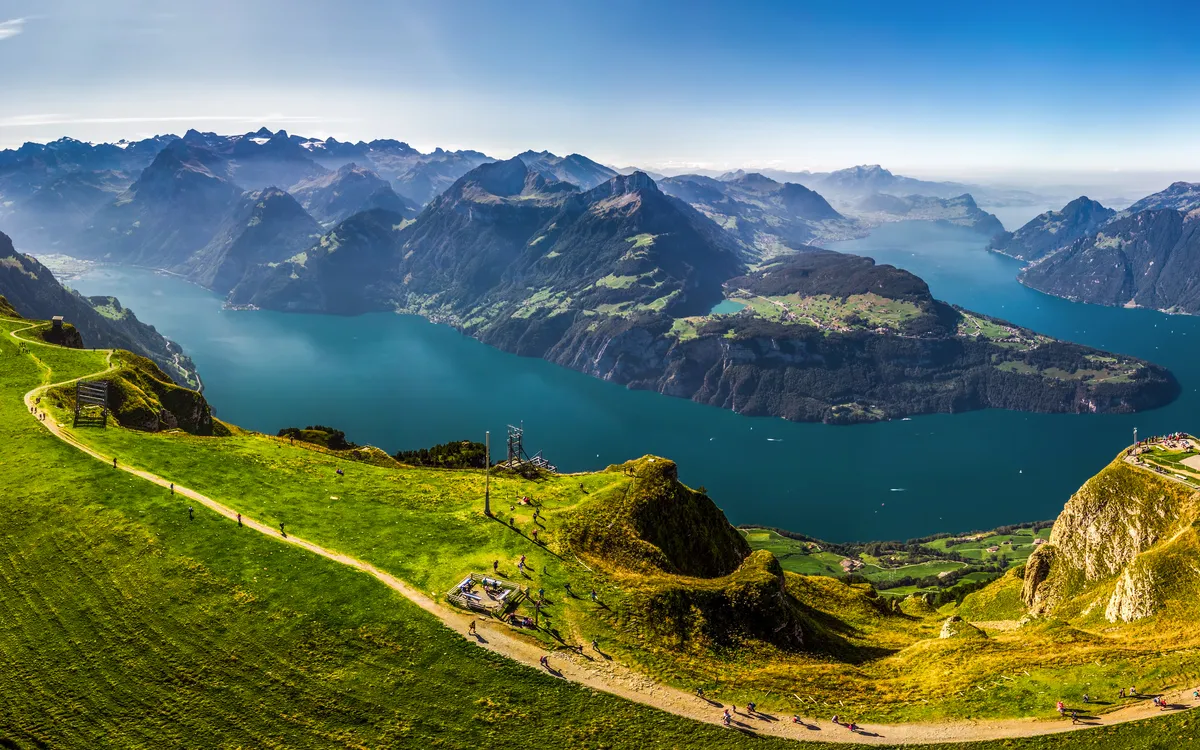 Bergpanorama mit See, grünen Wiesen und Wanderweg unter blauem Himmel.