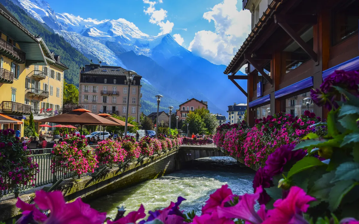 Blumen geschmückter Fluss zwischen Gebäuden in einer Stadt mit Bergen im Hintergrund.