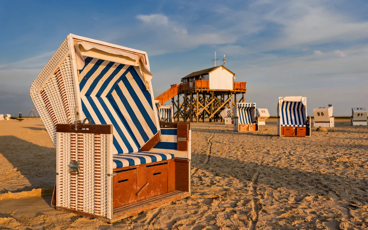Strandkorb am Sandstrand mit Pfahlbau im Hintergrund bei Sonnenuntergang.