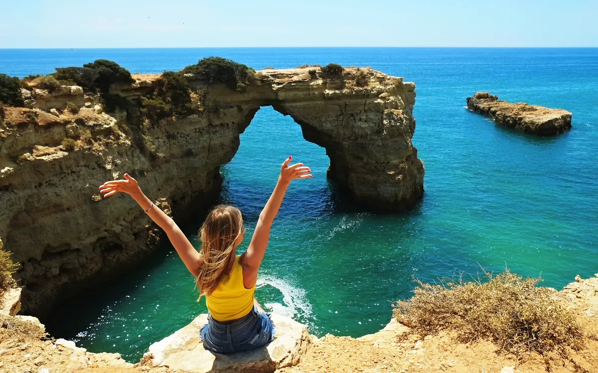 Frau sitzt auf Klippe mit Blick auf Felsformation und Meer.