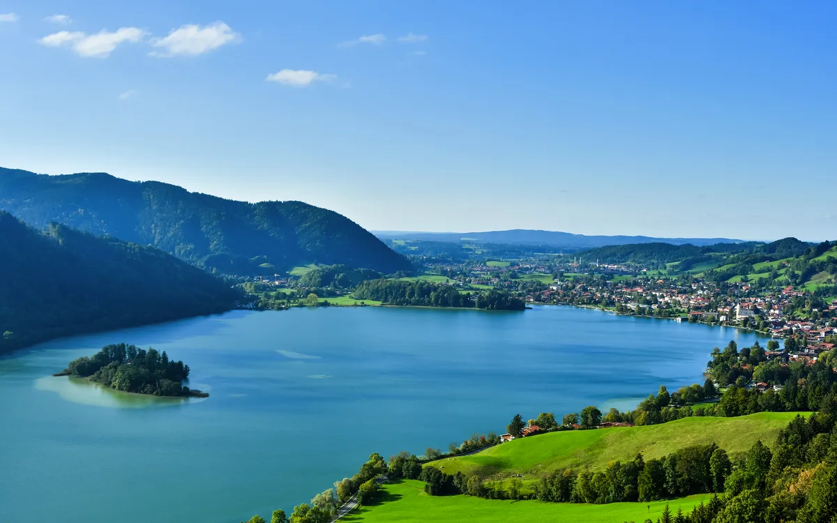 Blick auf einen See mit bewaldeten Hügeln und einer Stadt im Hintergrund.