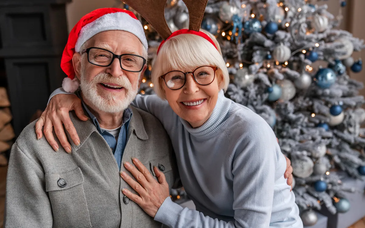 Älteres Paar mit Weihnachten Hüten lächelt vor geschmücktem Weihnachtsbaum.