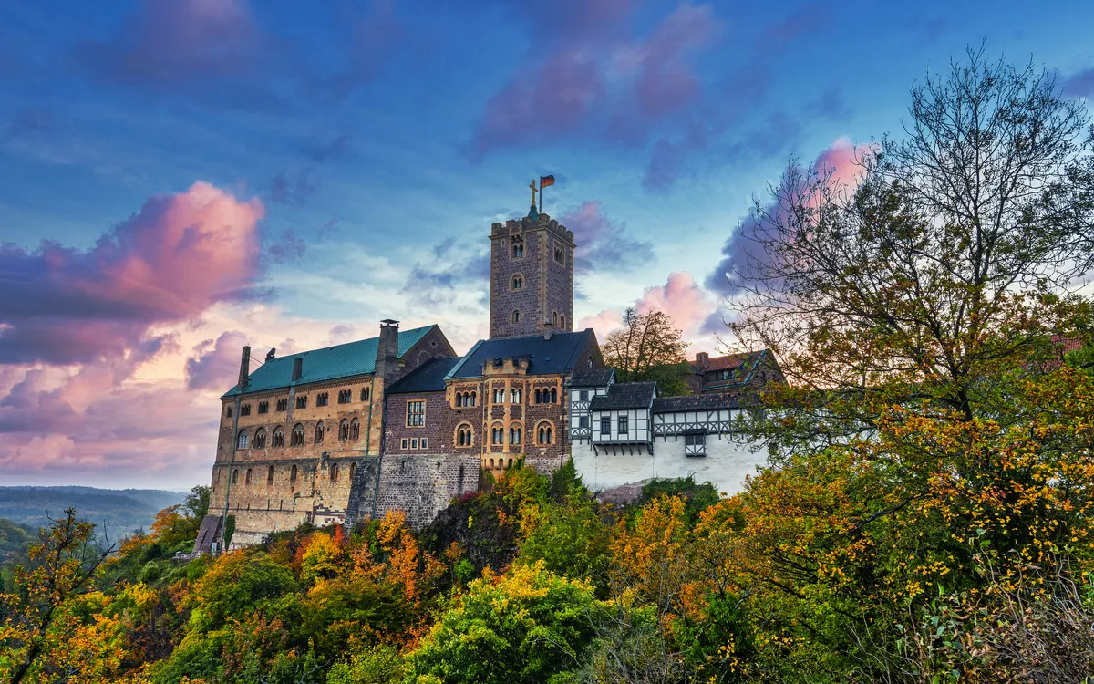 Burg auf einem Hügel bei Sonnenuntergang, umgeben von Herbstbäumen.