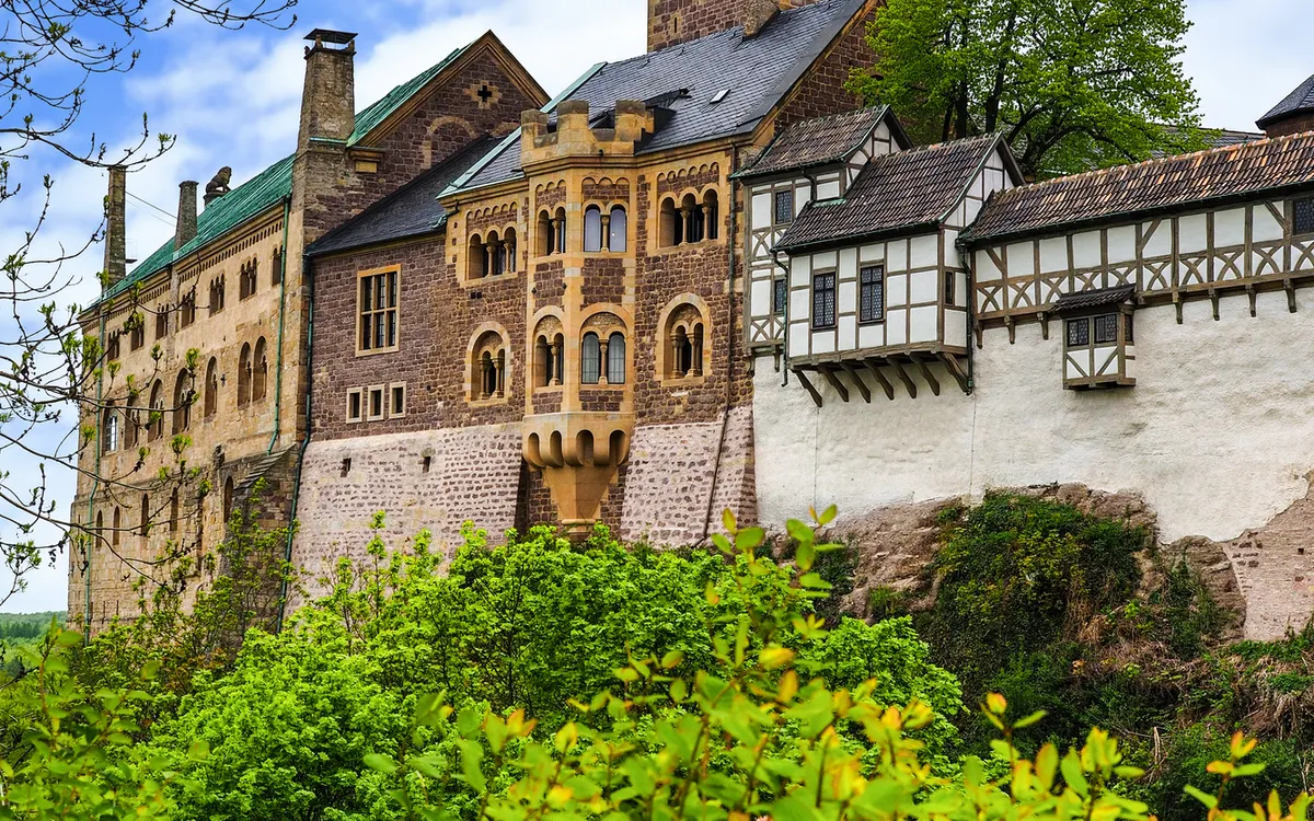 Historische Burg mit Turm und Fachwerkfassade, umgeben von grüner Vegetation.