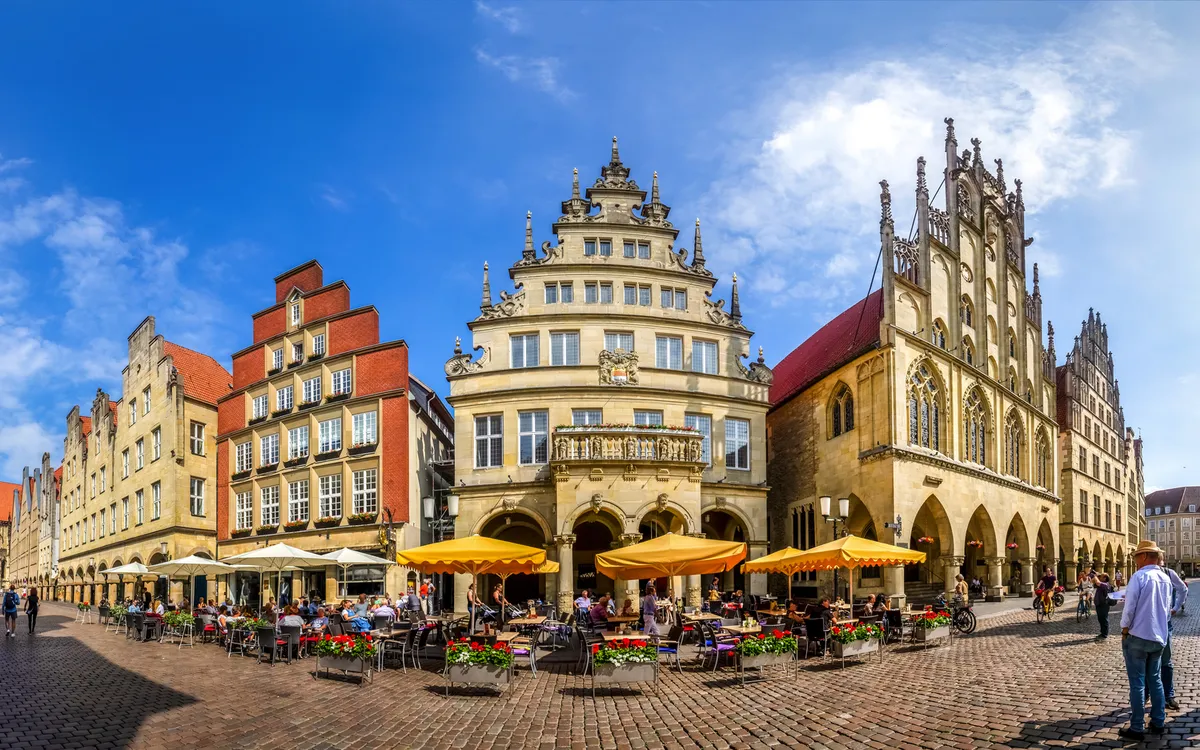 Historische Gebäude an einem belebten Platz mit Marktständen und Menschen unter blauem Himmel.