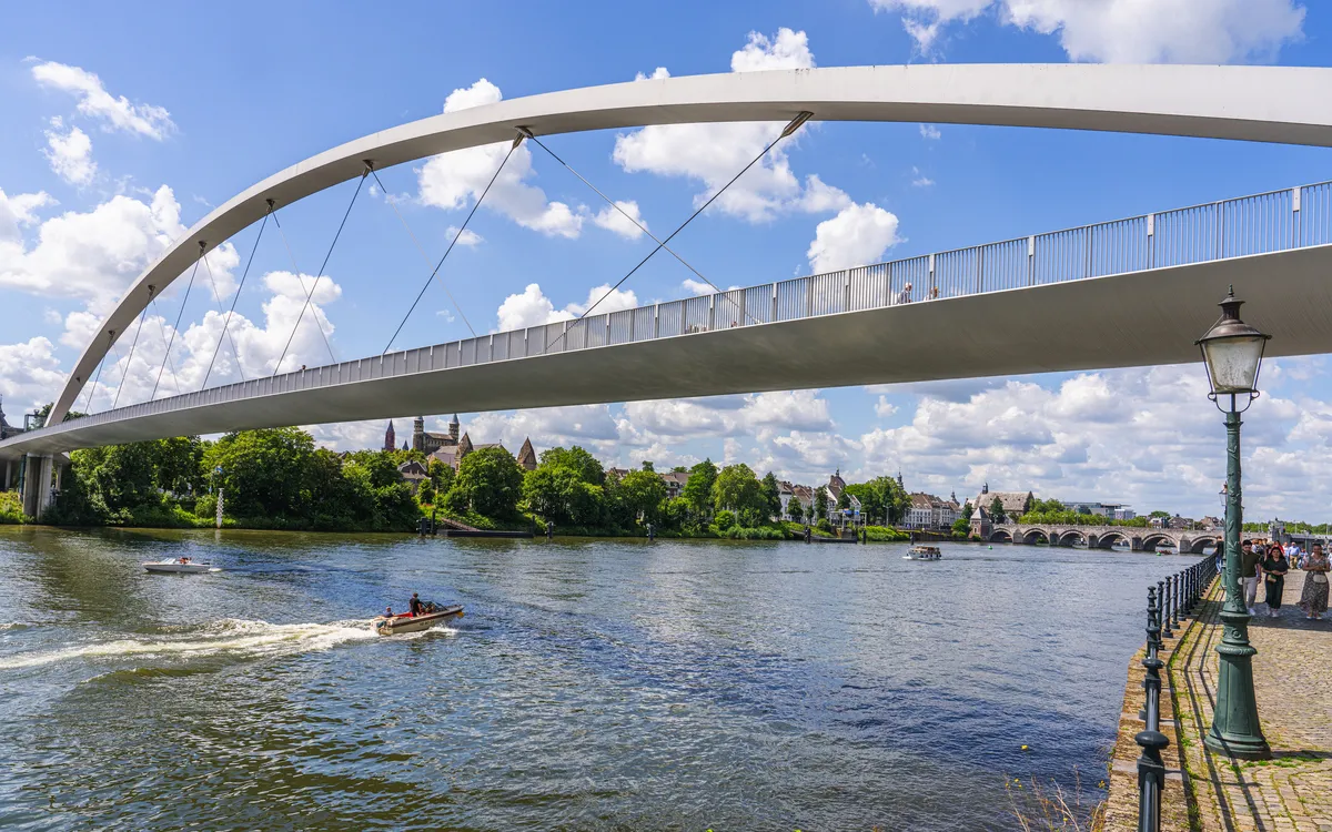 Brücke über einen Fluss mit Booten und Uferpromenade bei sonnigem Wetter.