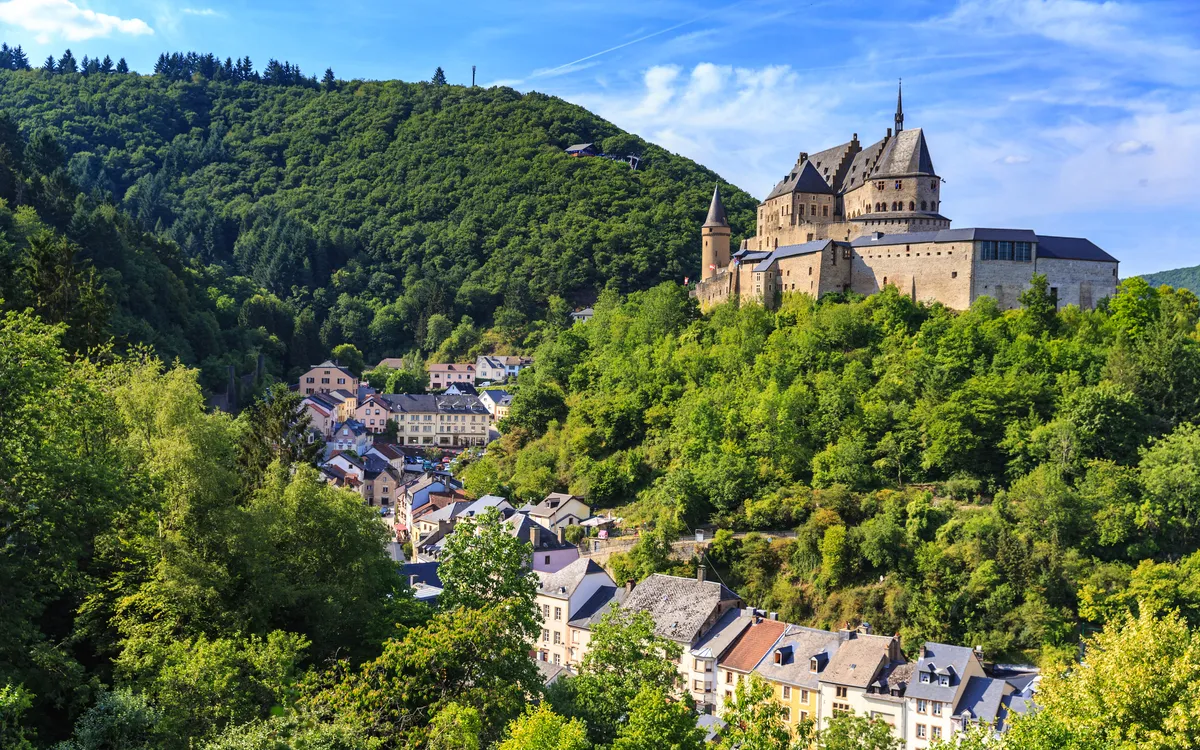 Burg auf Hügel mit grünen Wäldern und Dorf im Tal bei sonnigem Himmel.