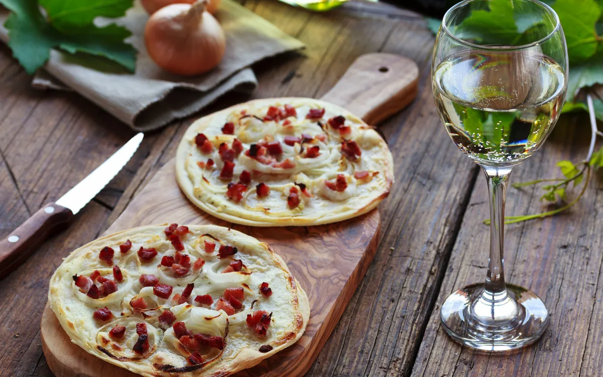 Zwei Flammkuchen auf Holzbrett mit Speck und Zwiebeln, daneben ein Glas Weißwein.